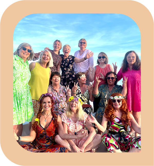 Gruppebilde av 13 damer i fargerike kjoler. Noen har blomsterkrans i håret og noen har solbriller på. Smiler i solen. Foto.