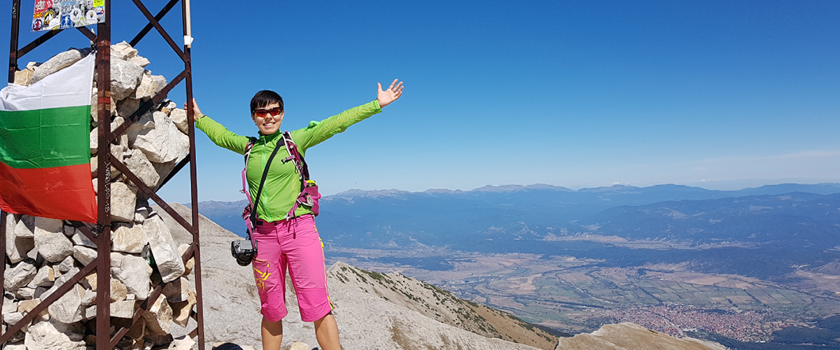 Norsk kvinne på toppen av Mount Vihren, rosa shorts og grønn topp, smiler med utsikt bak seg