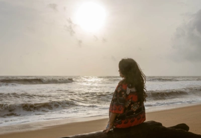 Kvinne som mediterer på en strand med ryggen til, vendt mot havet i mykt sollys og sandfargede toner. Foto.