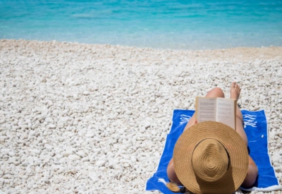 Kvinne med solhatt, ligger på hvit strand i Kroatia og leser en bok. Det klare turkise havet i bakgrunnen