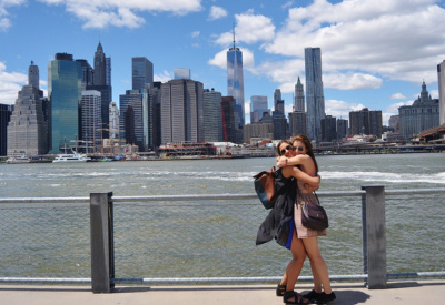 <p>To kvinner som står å klemmer hverandre ved Brooklyn Bridge. Utsikt over Manhatten og blå himmel med skyer</p>