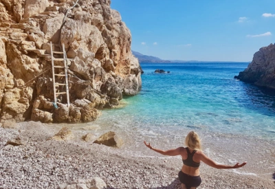 Foto av kvinne som nyter stranden i Hellas Karpathos. Hun sitter i sanden med armene ut. Vannet er turkist og himmelen blå.