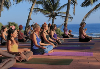 En gruppe mennesker sitter på yogamatter ute, utenfor AyurSoma i Kerala i India, med palmer og hav i bakgrunnen. Foto.