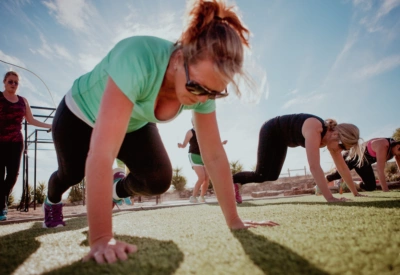 Nordisk kvinne på Mallorca i sort tights og grønn t-skjorte, utfører øvelsen Mountain climbers på gressplen, himmelen er blå. Hun har solbriller og hestehale.