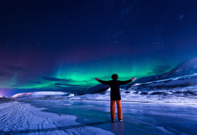 En person står med ryggen til og armene strukket ut, ute i snø og is, polarlandskap på Svalbard. Himmelen er mørkeblå med klart, grønt nordlys i hosrisonten. Foto.