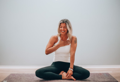 Yogalærer, Jannice Strand, sitter på en yogamatte. Smiler med øynene igjen og holder ene hånden på brystet. Foto.