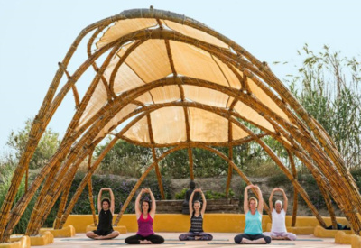 Utendørs yogashala laget av buet treverk og hvit seilduk. 5 yogadeltakere sitter i skredderstilling og utfører skulderstrekk på, det er fint vær, og sommer, deltakerne har på seg singlet og T-skjorte 