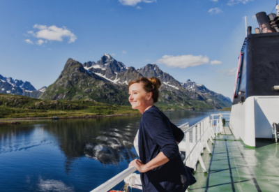Bilde av kvinne på dekke på Hurtigruten. Hun skuer utover havet med fjelltopper i bakgrunnen