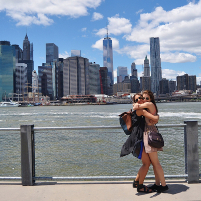 To kvinner som står å klemmer hverandre ved Brooklyn Bridge. Utsikt over Manhatten og blå himmel med skyer