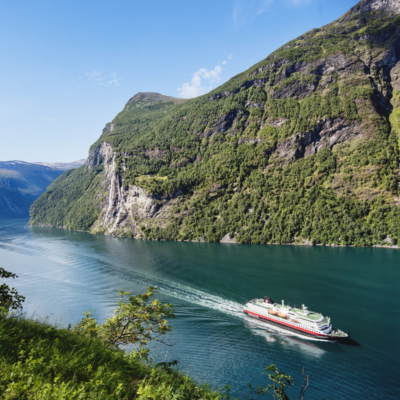Hurtigruten som seiler inn en blågrønn fjordarm med bratte fjell på siden. Foto.
