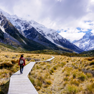 Kvinne som går på tur på 'Hooker Valley Track' på en plankesti innover mot bratte fjell med snødryss på toppene i Mount Cook National Park i New Zealand. Foto.