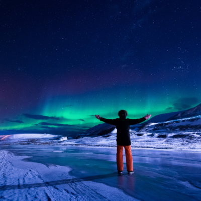 En person står med ryggen til og armene strukket ut, ute i snø og is, polarlandskap på Svalbard. Himmelen er mørkeblå med klart, grønt nordlys i hosrisonten. Foto.