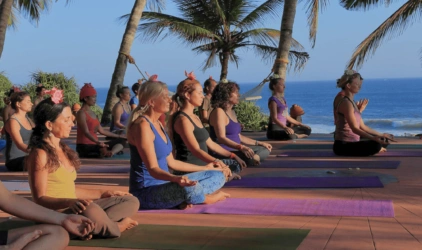 En gruppe mennesker sitter på yogamatter ute, utenfor AyurSoma i Kerala i India, med palmer og hav i bakgrunnen. Foto.