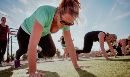 Nordisk kvinne på Mallorca i sort tights og grønn t-skjorte, utfører øvelsen Mountain climbers på gressplen, himmelen er blå. Hun har solbriller og hestehale.