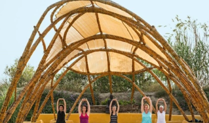 Utendørs yogashala laget av buet treverk og hvit seilduk. 5 yogadeltakere sitter i skredderstilling og utfører skulderstrekk på, det er fint vær, og sommer, deltakerne har på seg singlet og T-skjorte 