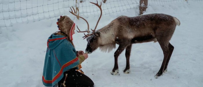 Samisk kvinne med tradisjonell samekofte og reinsdyr i snøen.