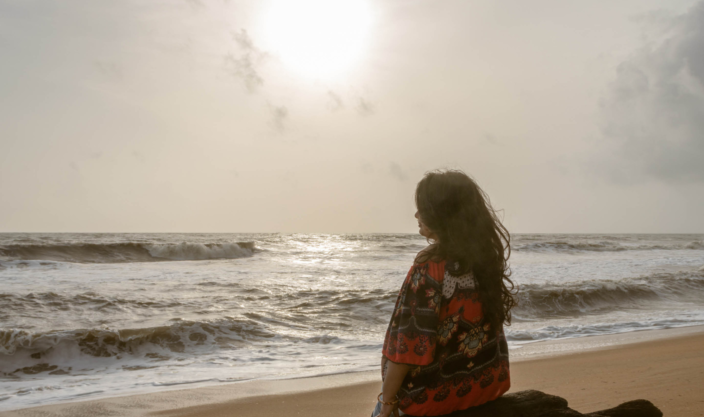 Kvinne som mediterer på en strand med ryggen til, vendt mot havet i mykt sollys og sandfargede toner. Foto.