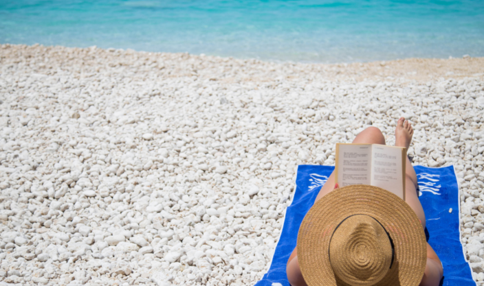 Kvinne med solhatt, ligger på hvit strand i Kroatia og leser en bok. Det klare turkise havet i bakgrunnen