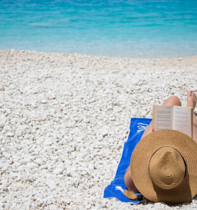 Kvinne med solhatt, ligger på hvit strand i Kroatia og leser en bok. Det klare turkise havet i bakgrunnen