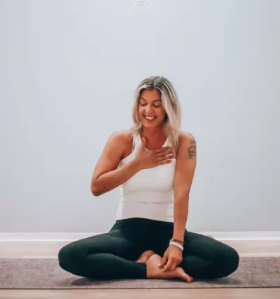 Yogalærer, Jannice Strand, sitter på en yogamatte. Smiler med øynene igjen og holder ene hånden på brystet. Foto.