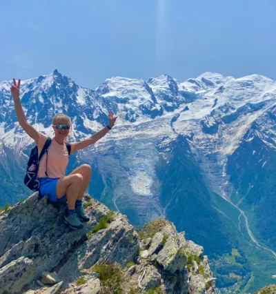 Kvinne (Kaisa Hafredal) sitter i shorts og t-skjorte på en fjelltopp med majestetisk utsikt til alpene i bakgrunnen. Foto.