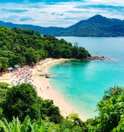 Bilde av en strand i Thailand. Turkist vann, gul sand og en frodig skog som omkranser stranden. Grønne åser i det fjerne.