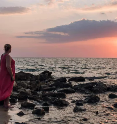 Kvinne i fjæra på sicilia, havet er blått, himmelen er rosa og det er solnedgang.