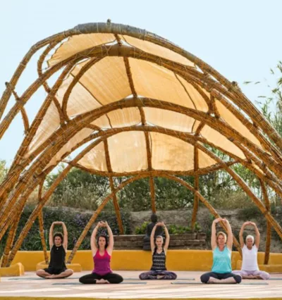 Utendørs yogashala laget av buet treverk og hvit seilduk. 5 yogadeltakere sitter i skredderstilling og utfører skulderstrekk på, det er fint vær, og sommer, deltakerne har på seg singlet og T-skjorte 