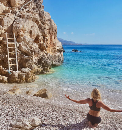 Foto av kvinne som nyter stranden i Hellas Karpathos. Hun sitter i sanden med armene ut. Vannet er turkist og himmelen blå.