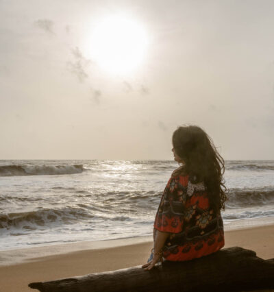 Kvinne som mediterer på en strand med ryggen til, vendt mot havet i mykt sollys og sandfargede toner. Foto.