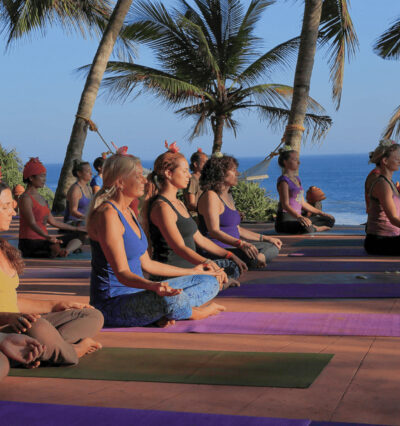 En gruppe mennesker sitter på yogamatter ute, utenfor AyurSoma i Kerala i India, med palmer og hav i bakgrunnen. Foto.